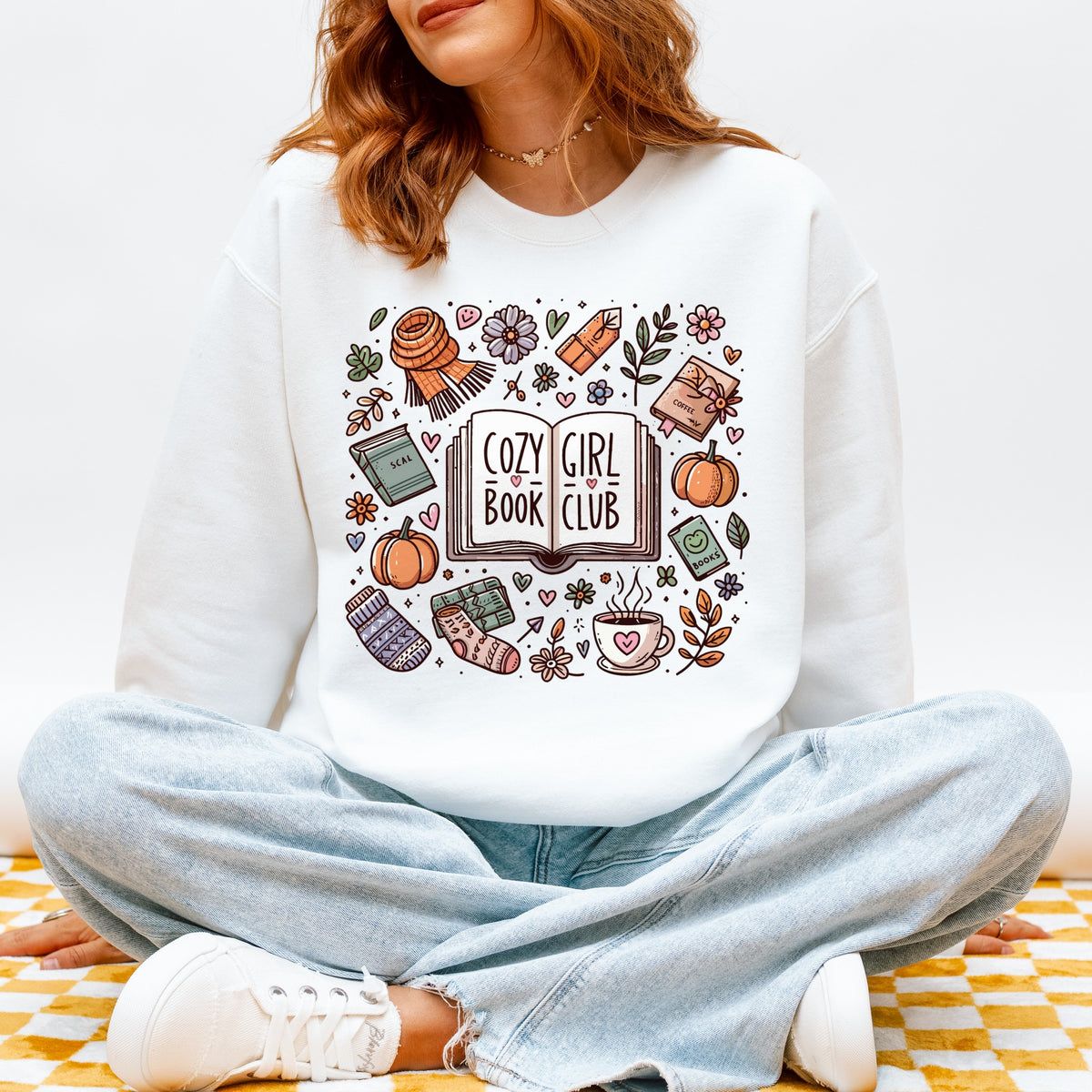 Woman wearing a white sweatshirt with an illustrated Cozy Girl Book Club design of books and coffee