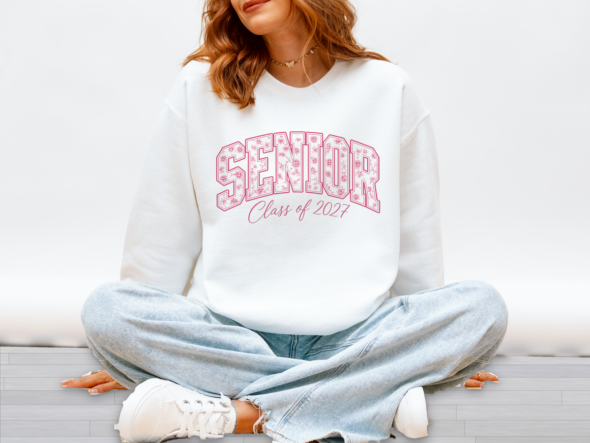 Woman sitting cross-legged in a white SENIOR Class of 2027 pink floral varsity sweatshirt